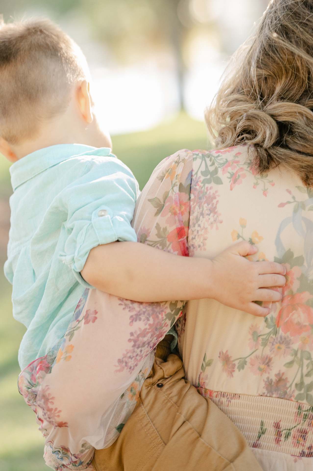 A young child wraps an arm around a woman's back as she holds them close, her floral dress soft in the afternoon light